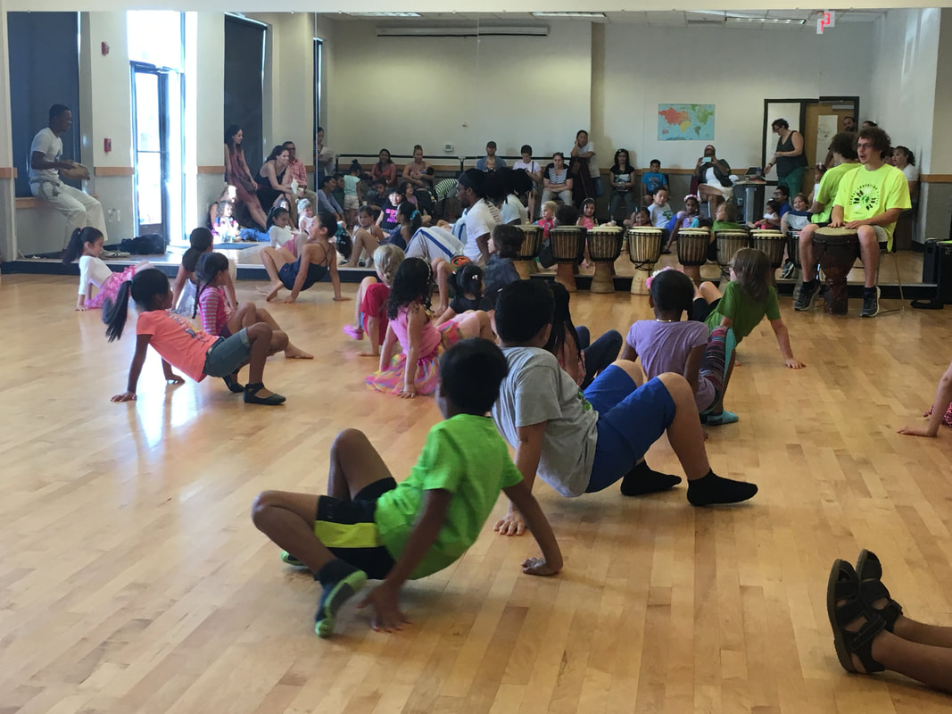 A dance studio of young children on the floor with one hand in the air practicing Capoeria with a teacher and traditional live music.