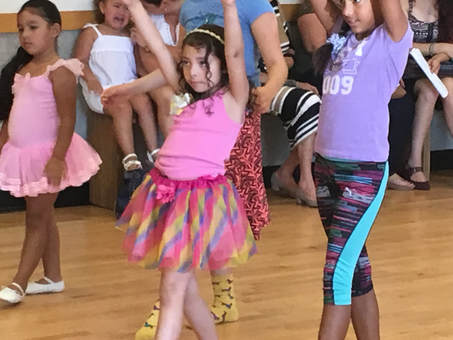 Three young girls, one in a pink tutu, another in a multicolor tutu and the other in workout clothing, are all practicing dance moves.