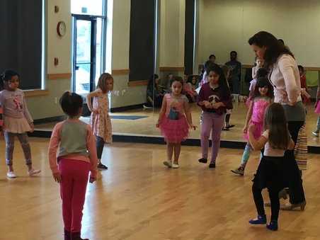  A circle of young girls in a dance studio are mimicking their dance teacher but tapping their toes as they learn tap dance.